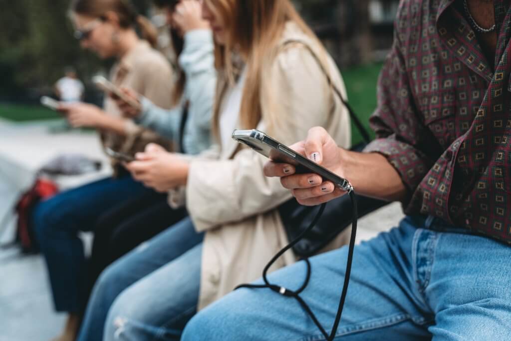 Four friends are using smart phones while they are sitting in a public park in the city. Detail of the hands holding the mobile phones.