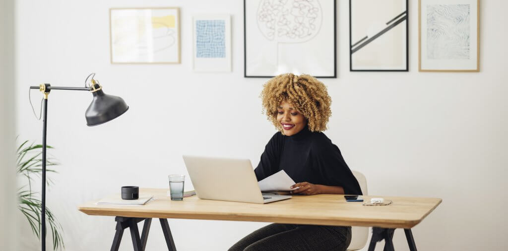 Cheerful and beautiful woman sitting at her desk. She is looking at something on her laptop while holding a paper in her hand. Her mobile phone, sunglasses and ear buds case are on the desk as well as a glass of water.