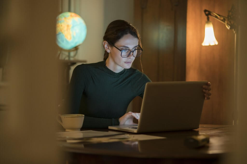 Stock photograph of a good looking, professional woman working in her home study at night with a laptop on the table.
