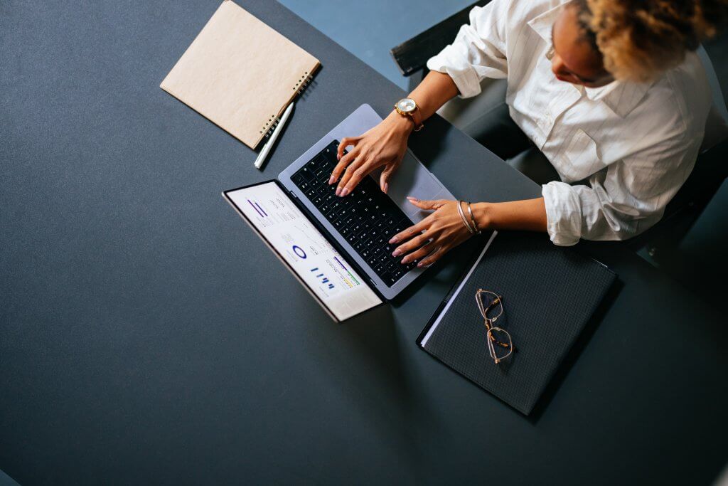 From above photo of an anonymous African-American woman analyzing business graph on a laptop computer while sitting at restaurant desk with notebook, pen and eyeglasses.