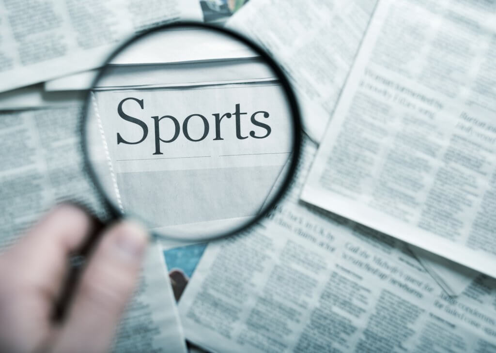 man holding magnifying glass on the "sports" word in the stack of newspapers, blue toned image