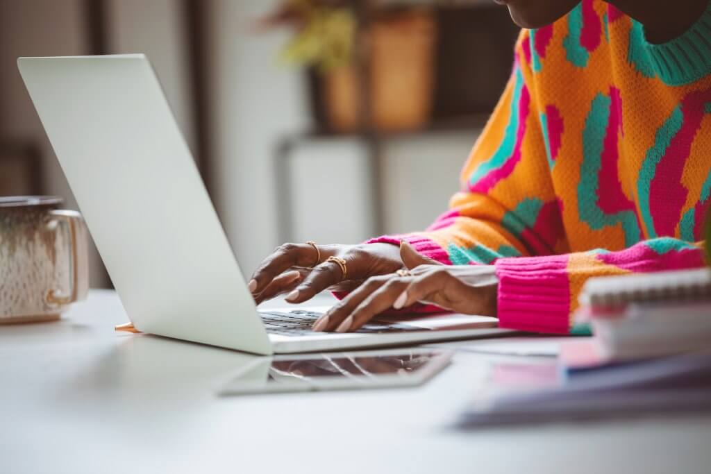 A young woman wearing colorful sweater sitting at the desk at home and using laptop. Close up of hands, unrecognizable person.