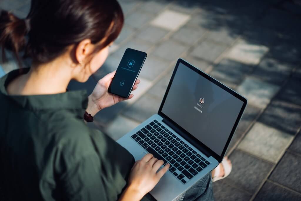 Young Asian businesswoman sitting on the bench in an urban park working outdoors, logging in to her laptop and holding smartphone on hand with a security key lock icon on the screen. Privacy protection, internet and mobile security concept